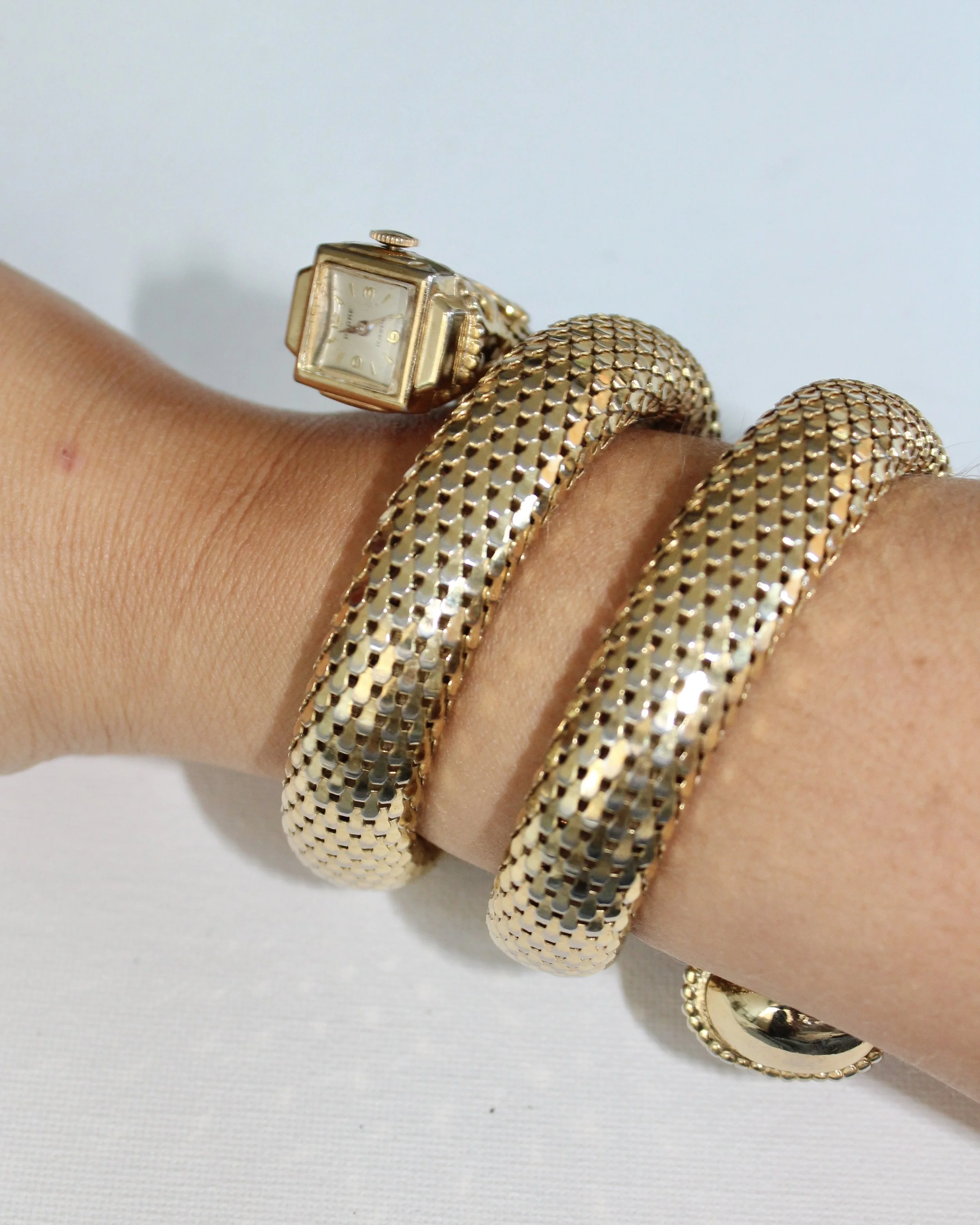 A hand wearing a gold watch and two gold mesh rings, with the rings wrapped around the wrist, against a plain white background.