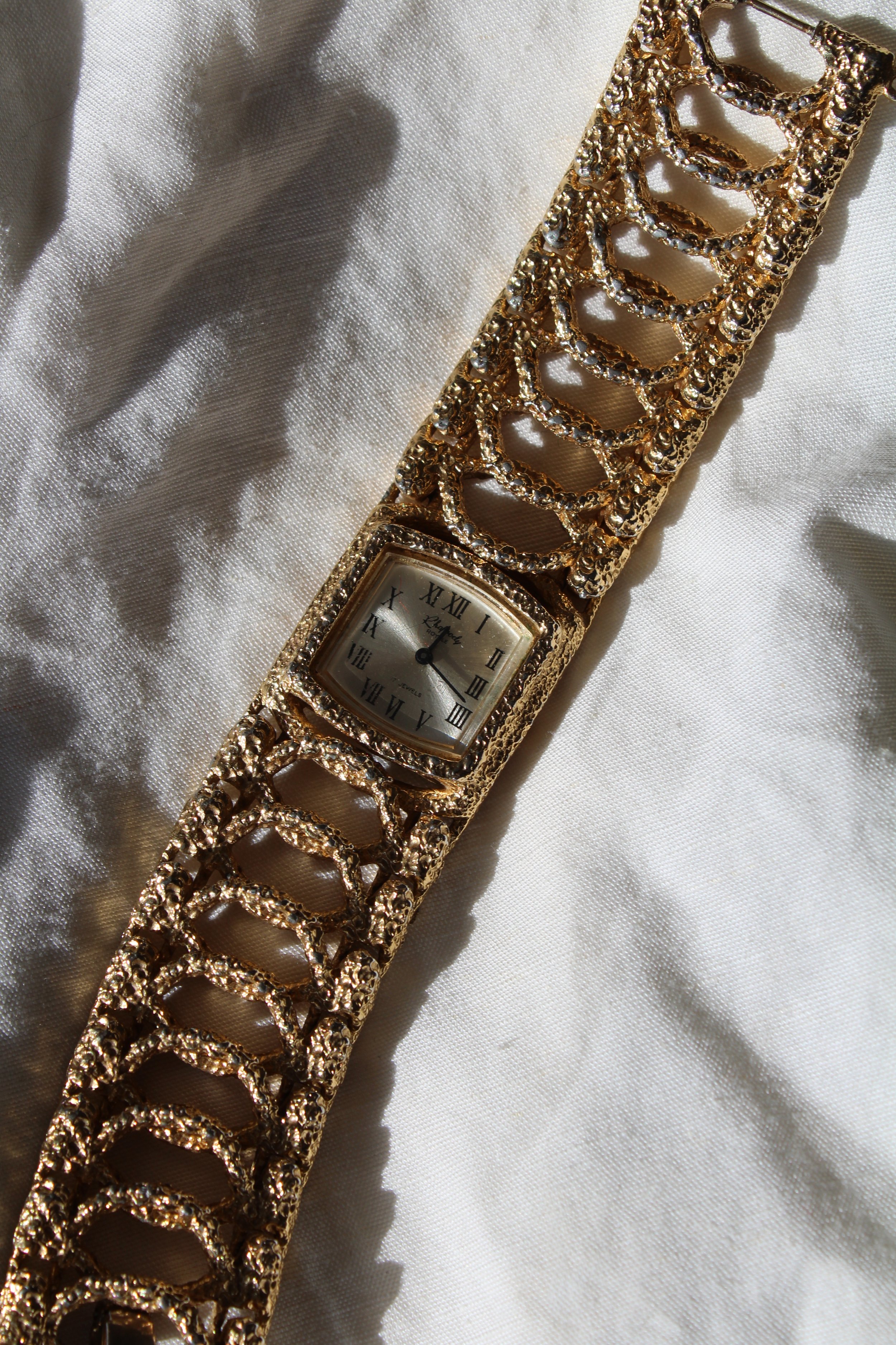 Gold wristwatch with a square face and Roman numerals on a sparkly, decorative band, placed on white satin fabric.