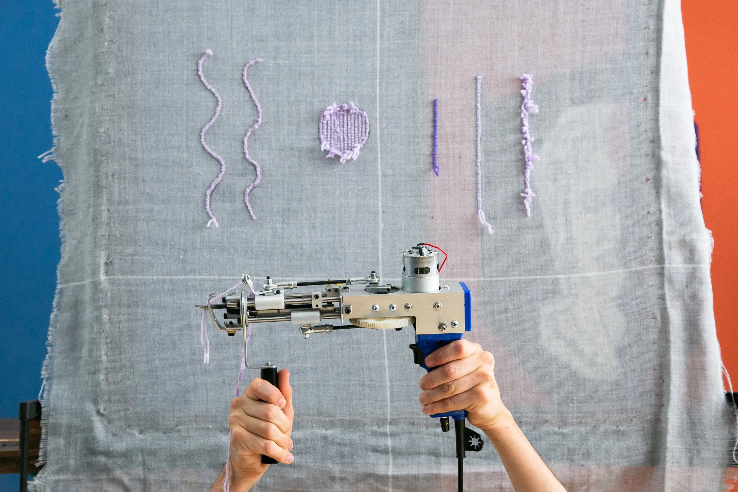 Main tenant un robot de couture ou de broderie pointant un tissu en toile, avec des fils de différentes couleurs et formes en arrière-plan.