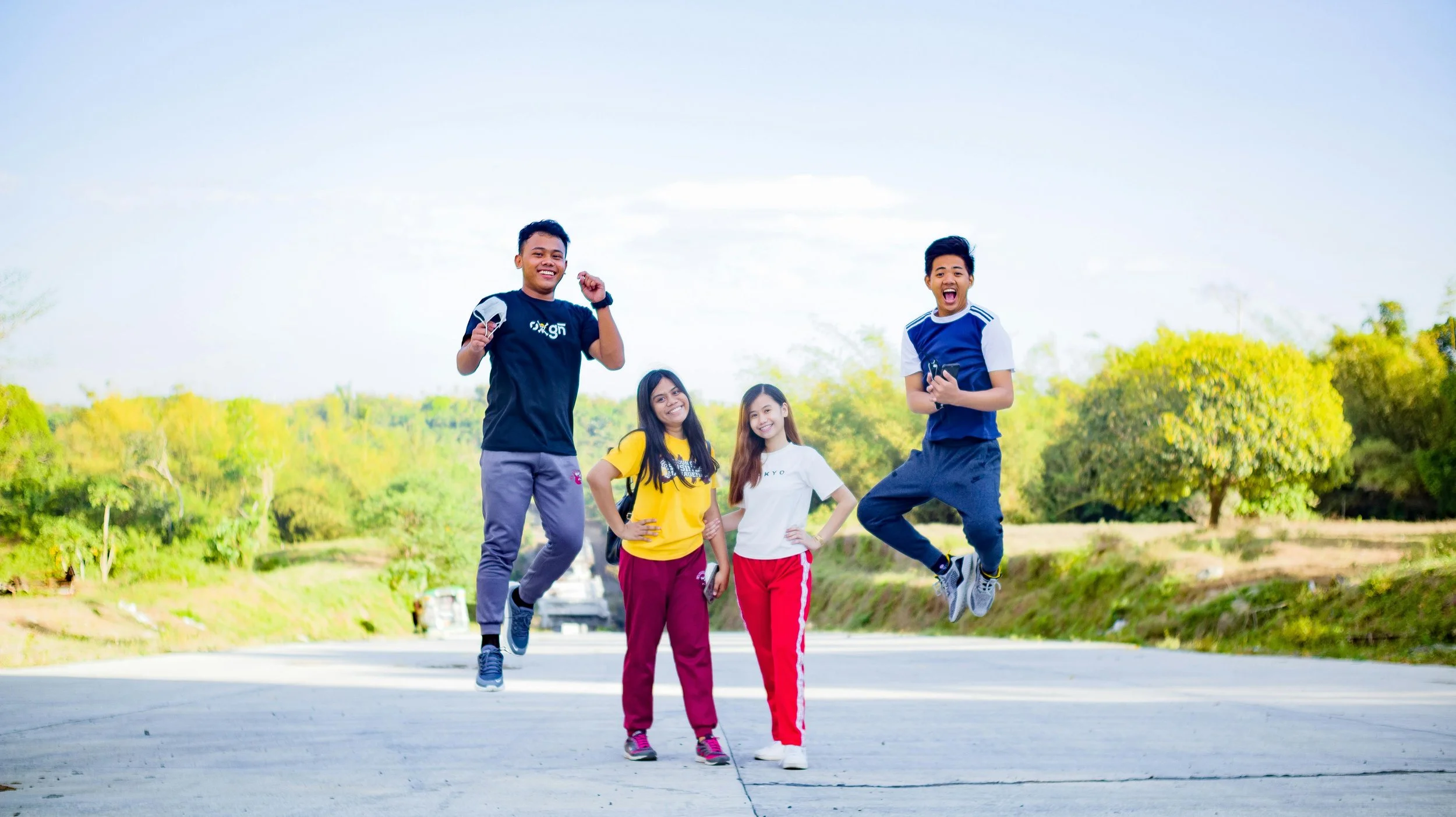 4 teenagers outside surrounded by trees, two teens jumping in the air