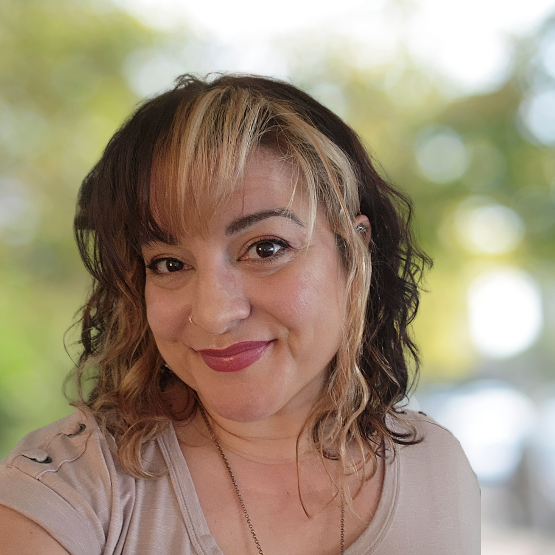 A woman with shoulder-length wavy hair, wearing a black lace top, smiling outdoors with a background of blurred green and white lights.