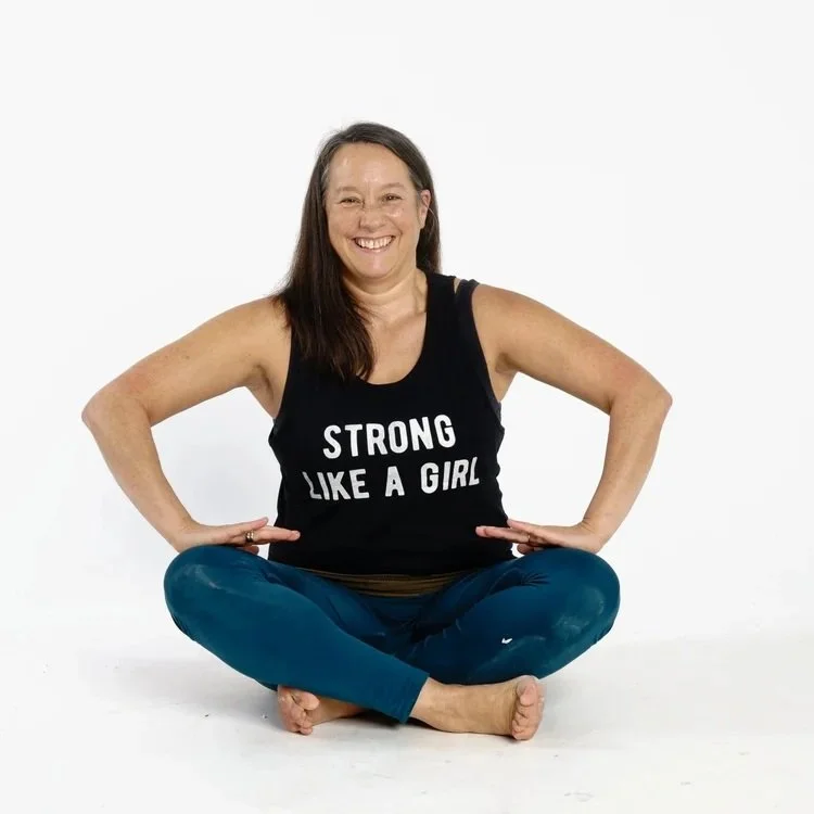 Abby sitting crossed legged in the hypopressive pose Hestia wearing a black vest top with the logo strong like a girl on the front and teal leggings and bare feet.