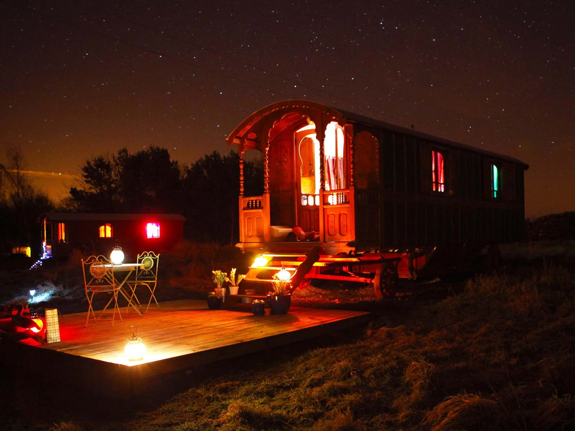An illuminated vintage wooden train car with open door and colorful window lights at night under a starry sky, displayed on a platform with outdoor furniture and lanterns.