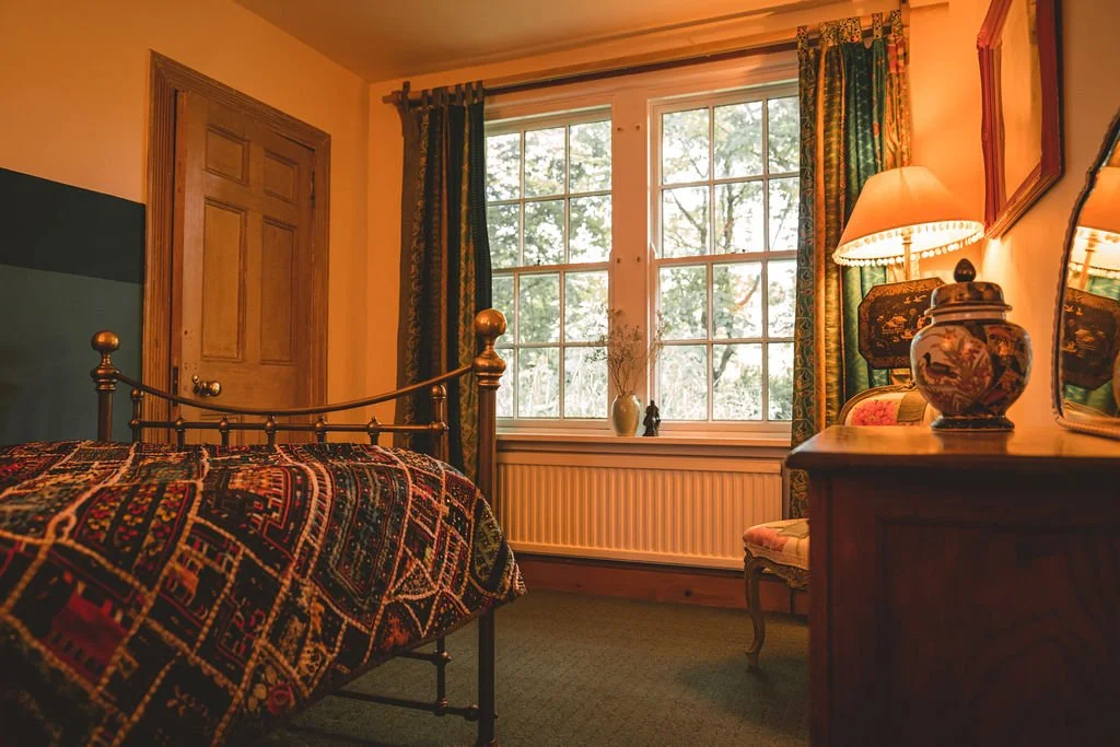 A cozy bedroom with a bed, a window with curtains, a nightstand with a lamp and decorative vase, and a wooden door.