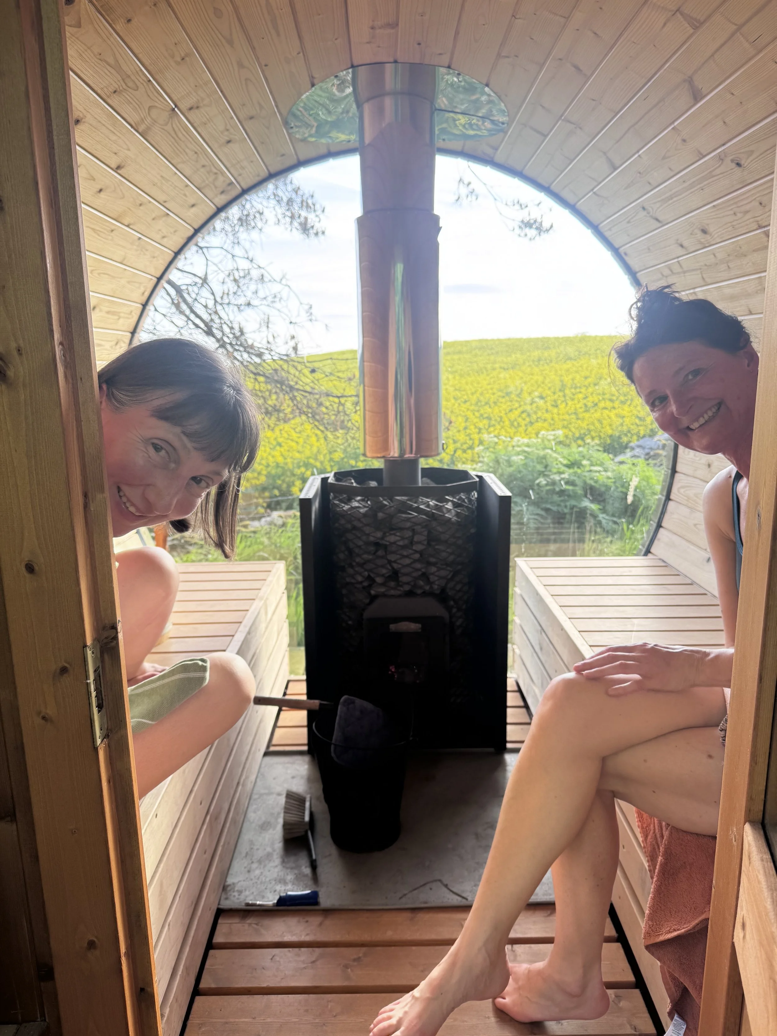 Two roulotte clients in a sauna wood fired enjoying the meadow beyond and smiling at the camera