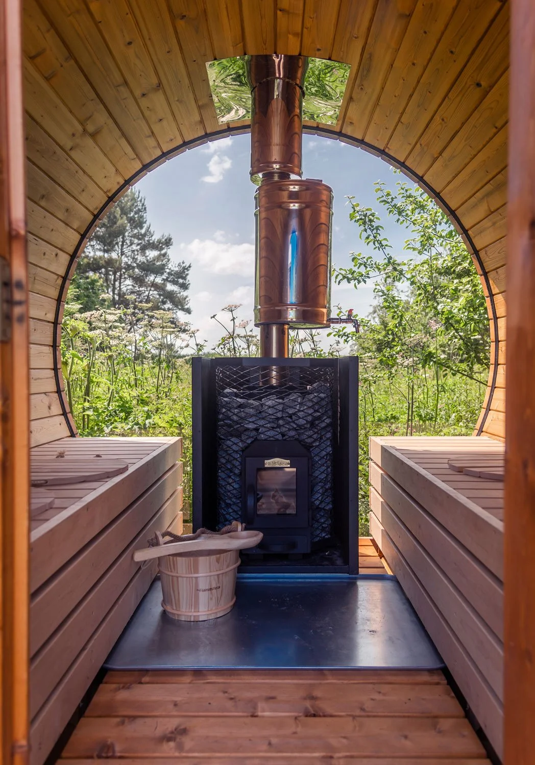 Interior of a wooden sauna with benches on both sides, a stove with a copper chimney, a bucket with a ladle, and lush greenery outside