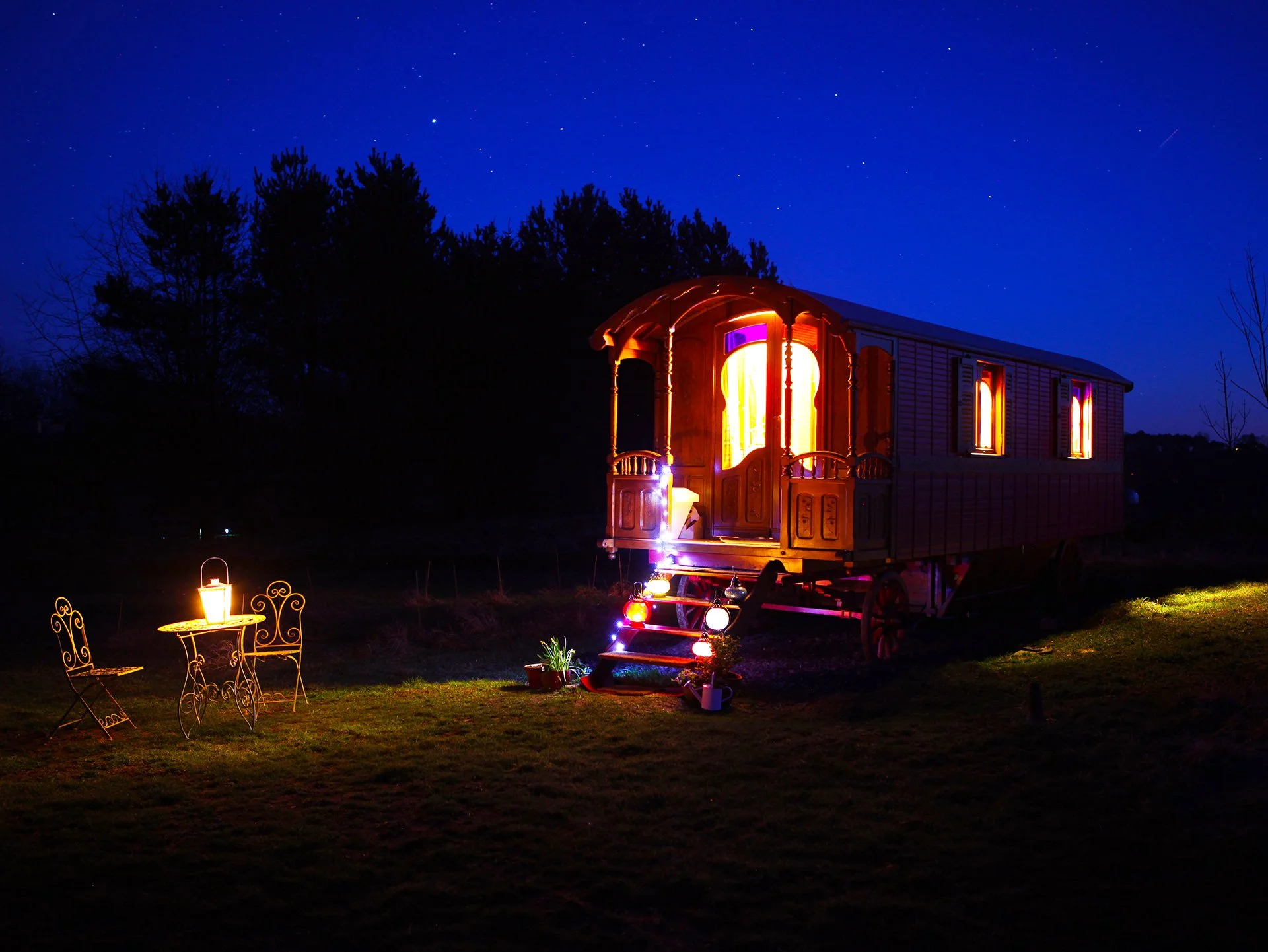 A brightly lit wooden caravan with windows and doors, located outdoors at night under a starry sky, with outdoor lighting and small garden decorations.