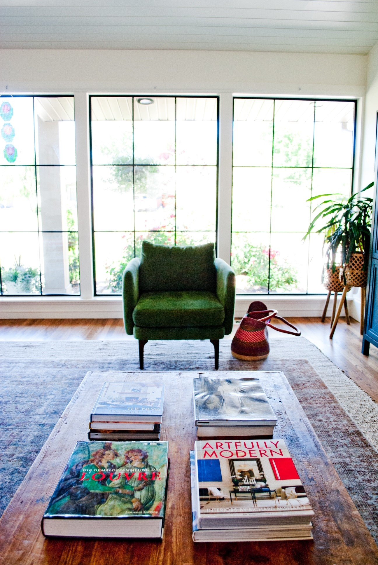 Interior living room with large windows, a green armchair, a wooden coffee table with books, a potted plant, and a woven basket, during daytime.