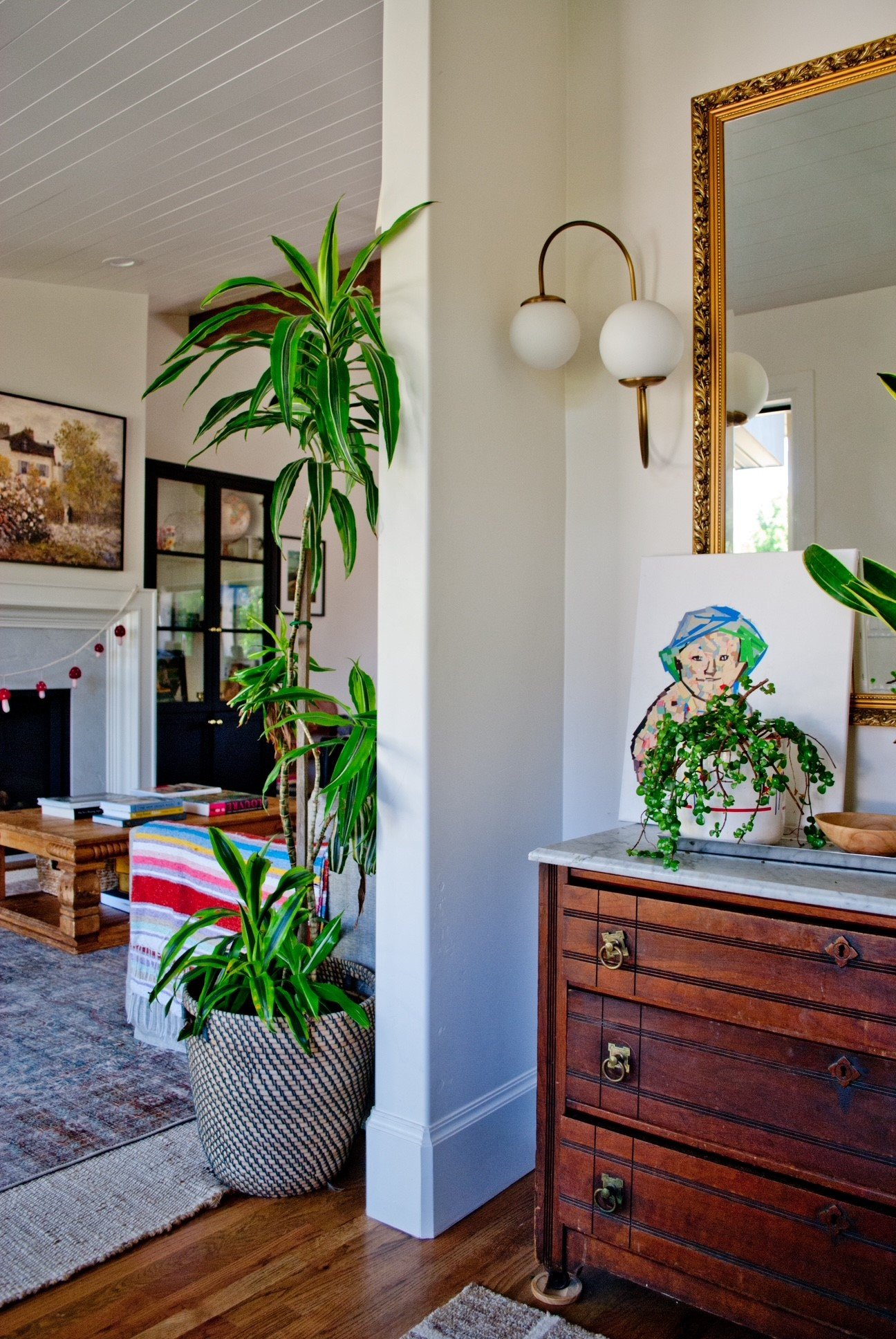 Interior view of a living room with a large potted plant, wooden sideboard, framed mirror, artwork, and a cozy fireplace.