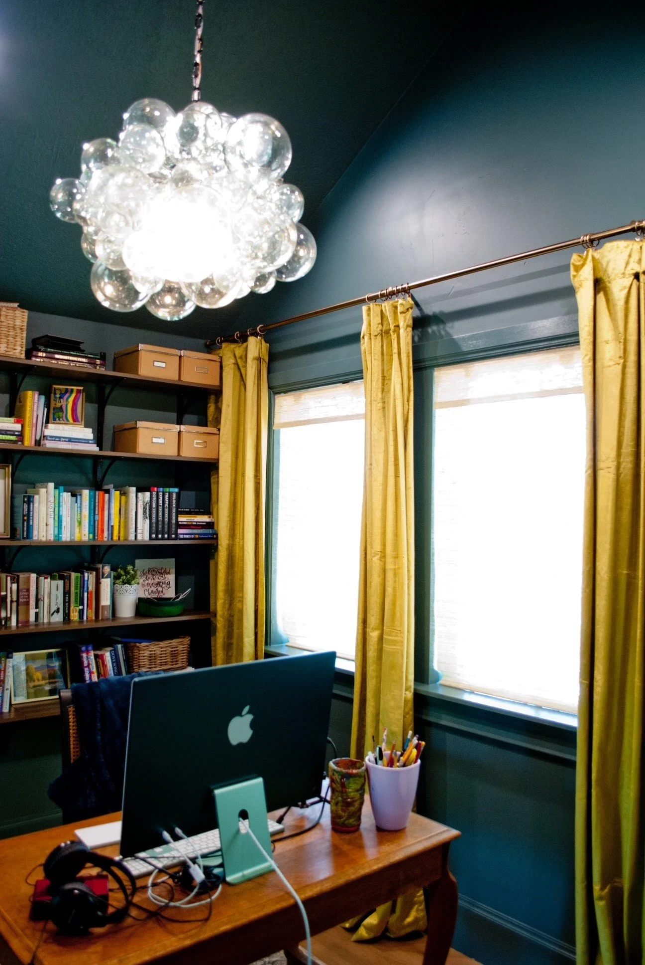 A home office with a wooden desk, a large monitor, and a collection of pens and art supplies in cups. Bright yellow curtains frame two windows, and a dark teal bookshelf holds books, boxes, and decorative items. A modern bubble chandelier hangs from 