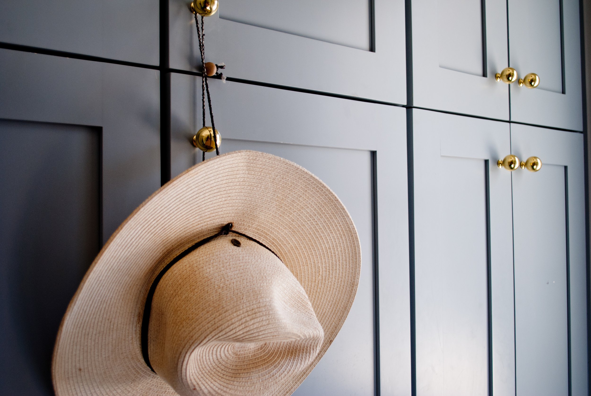 A beige wide-brimmed hat hanging on a black hook on a black paneled wall with gold knobs.