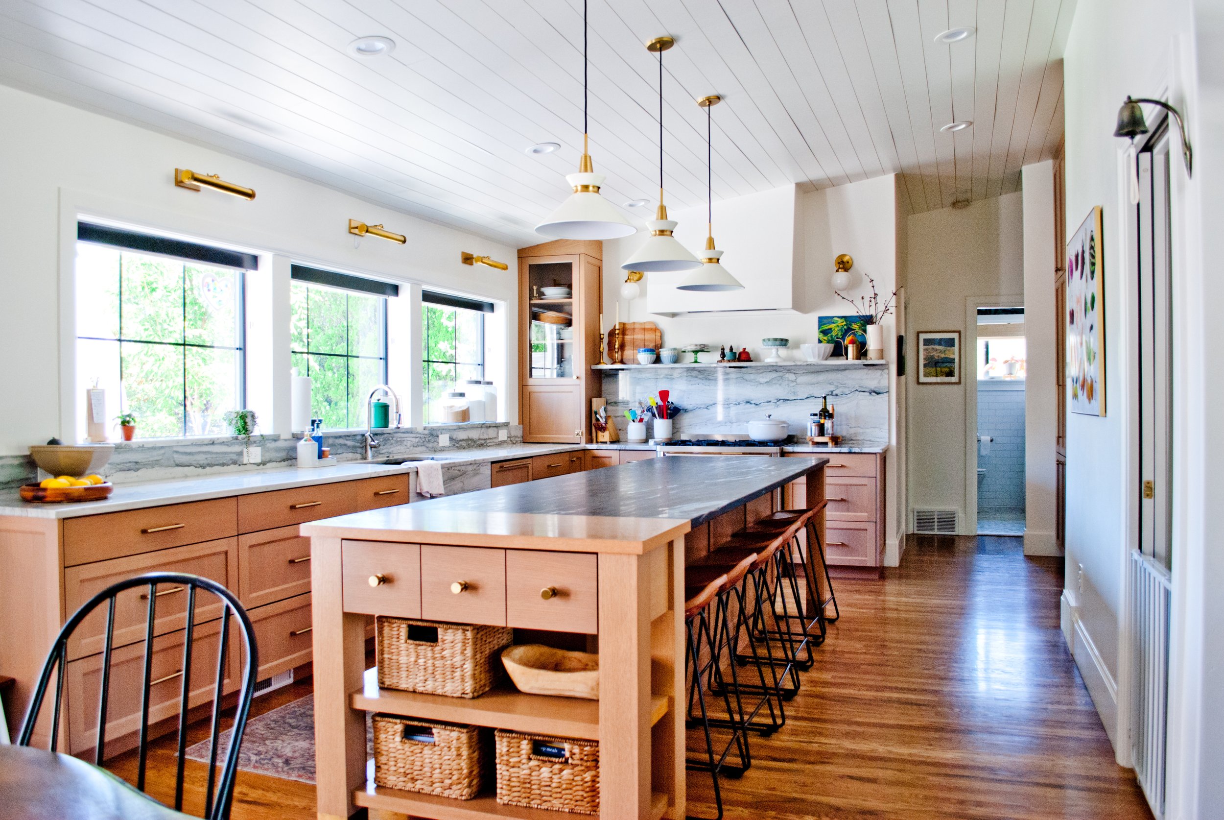 Bright kitchen with large windows, wooden cabinets, a marble backsplash, black countertops, and a center island with wooden drawers and baskets. Modern pendant lights hang from the ceiling, and bar stools are positioned along the island.