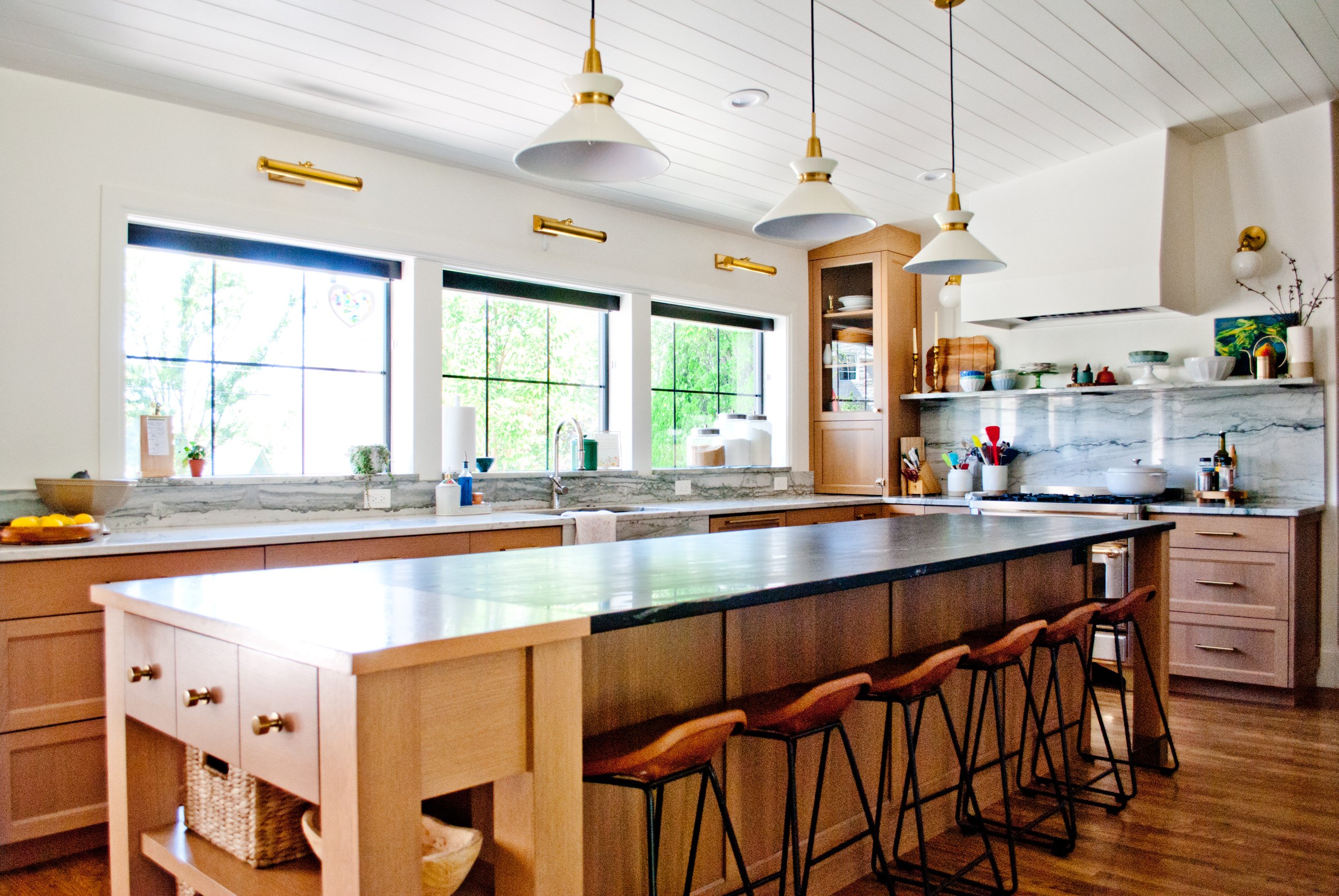 Bright kitchen with large windows, wooden cabinets, marble backsplash, a large island with seating, and gold-accented pendant lights.