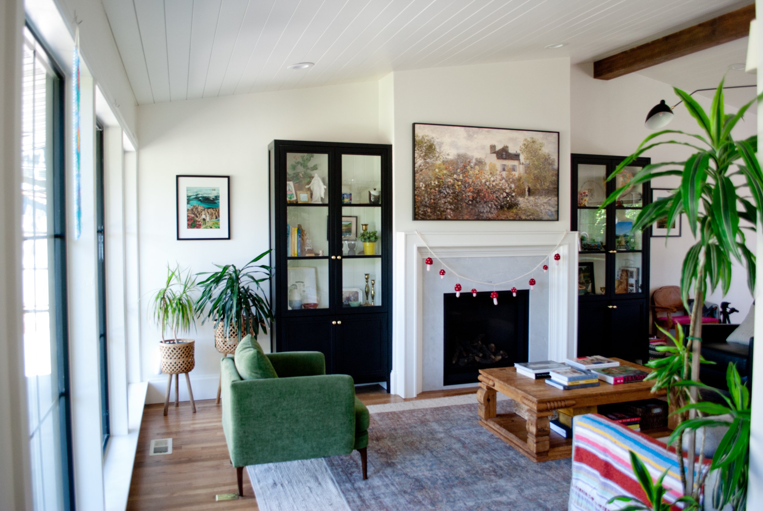 Living room with white walls, wooden flooring, a white fireplace with a garland, black display cabinets, green armchair, wooden coffee table, large plants, wall art, and a sliding glass door to the left letting in natural light.
