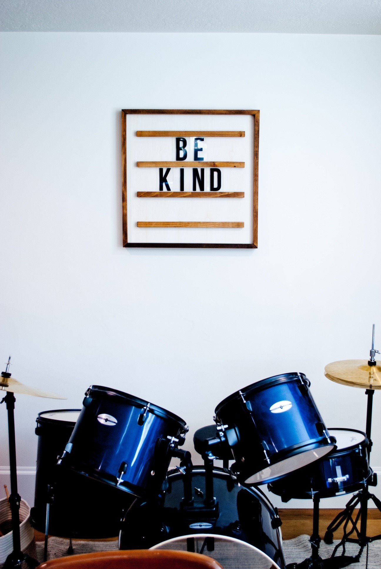 A room with a drum set and a white wall hanging that says 'BE KIND'.