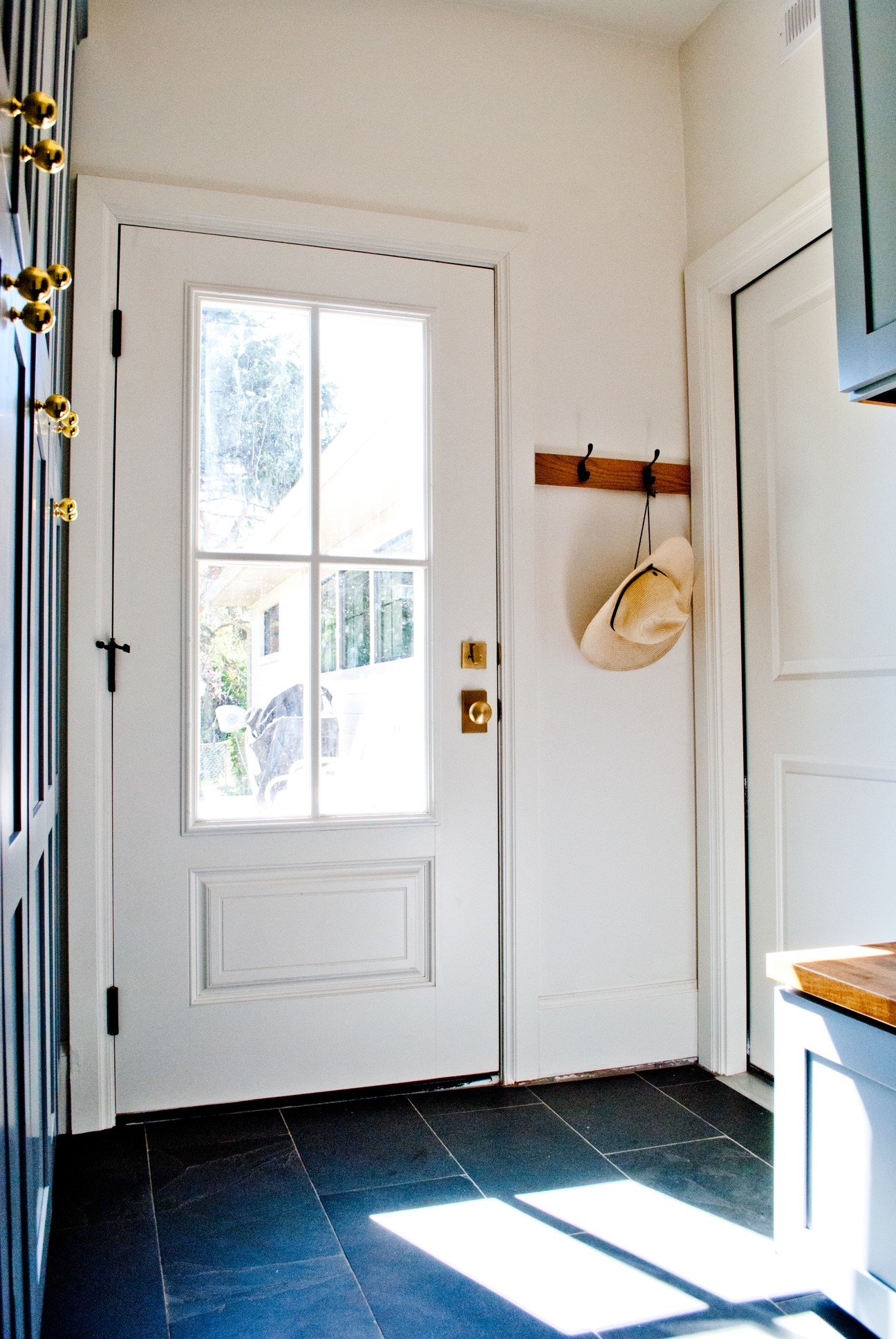 Interior view of a white door with glass panes leading outside, with a wooden coat rack on the wall holding a tan hat, sunlight streaming through the glass, and black tiled flooring.