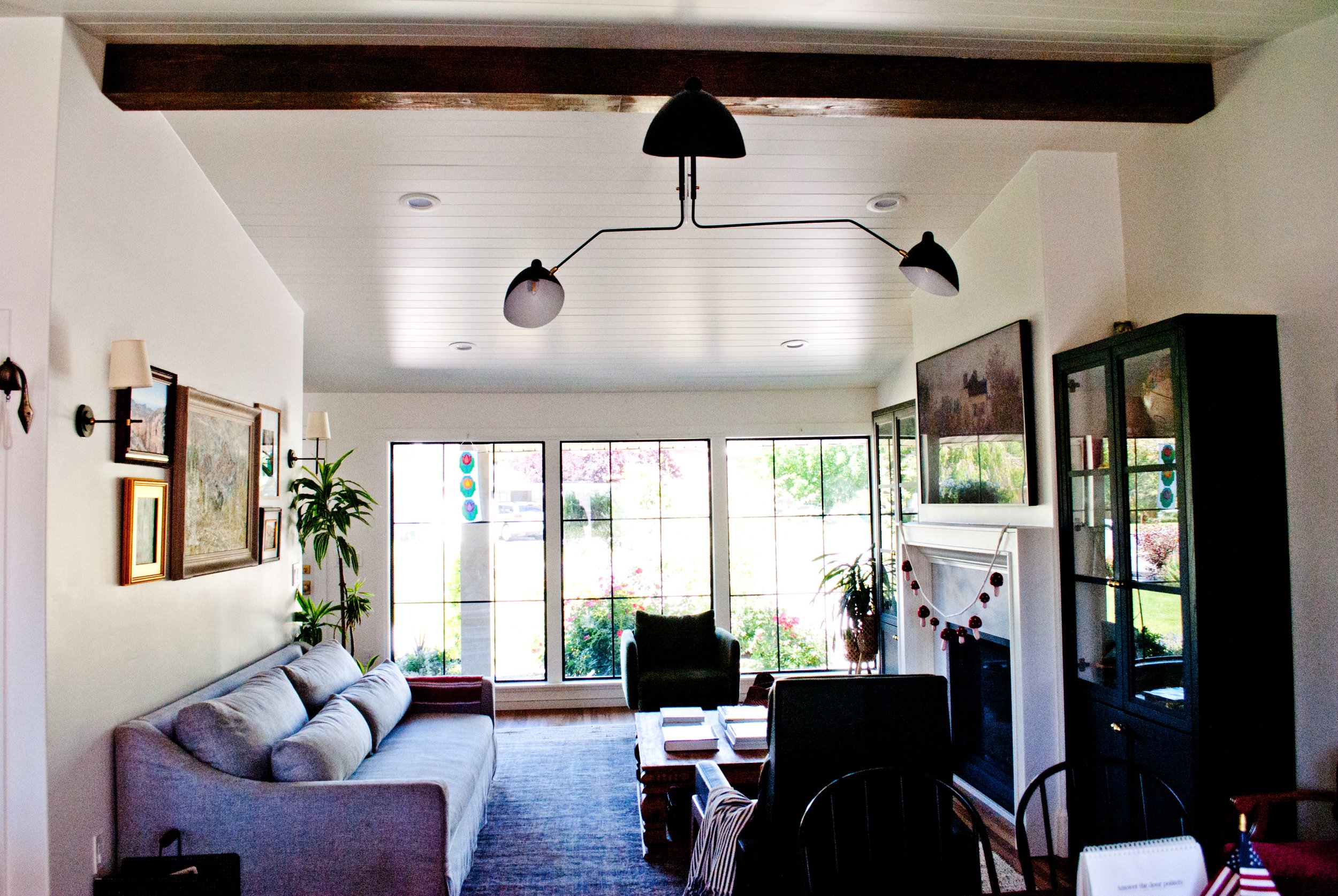 A living room with a gray sofa, black armchair, coffee table, and built-in fireplace. Large windows let in natural light, and there are decorative plants, framed artwork, and a modern chandelier hanging from the ceiling.