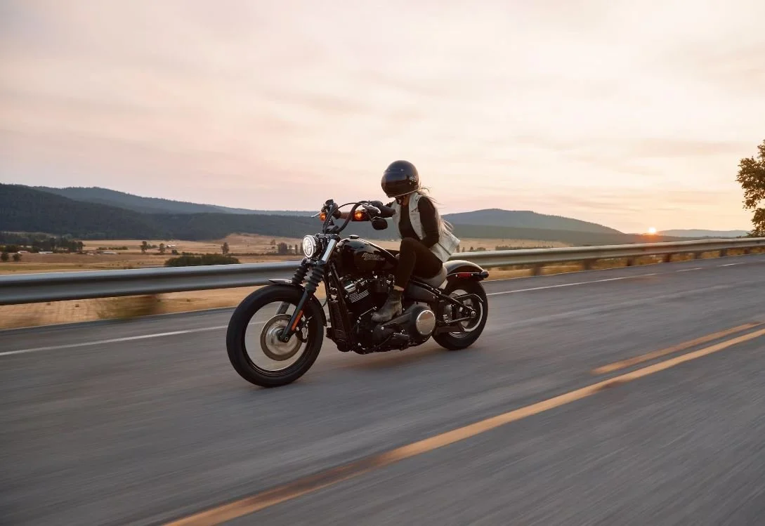 A woman wearing a helmet riding a motorcycle on an open road during sunset, with mountains and trees in the background.