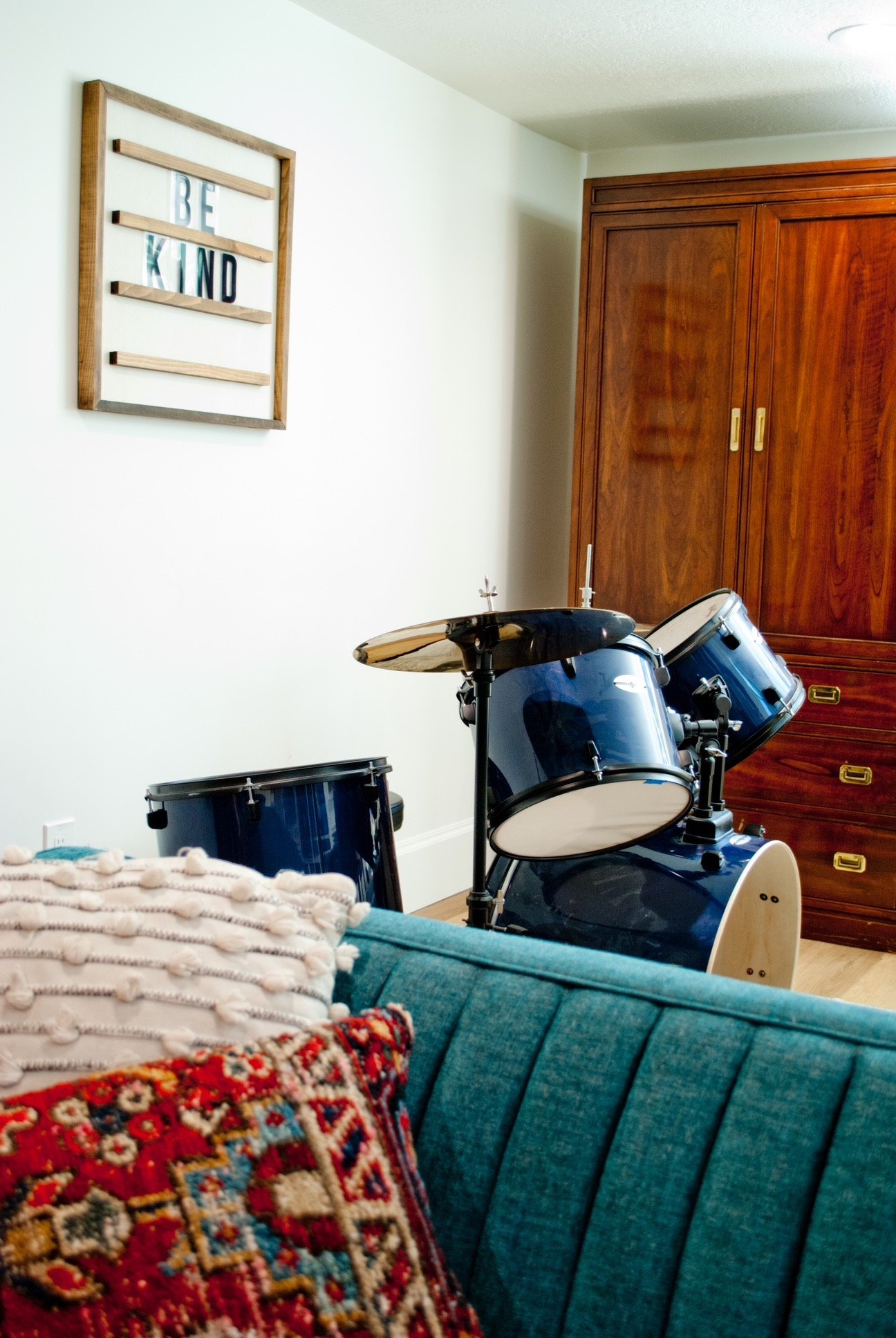 A blue drum set with cymbals in a room next to a wooden wardrobe and a white wall with a framed decorative sign that reads 'BE KIND'. There are colorful cushions on a teal sofa in the foreground.