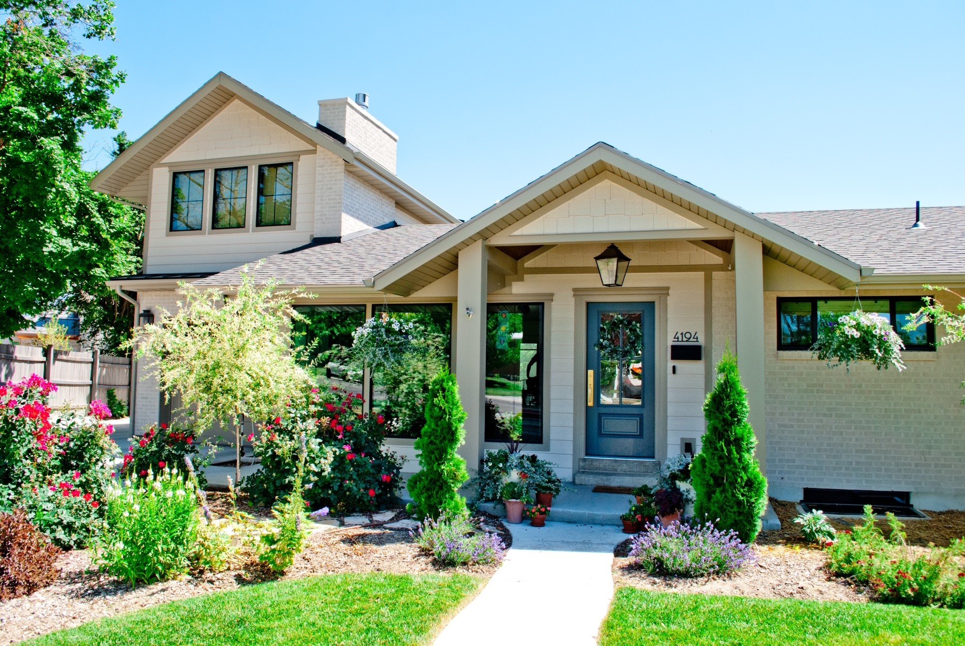 A modern house with a garden, featuring a white exterior, a porch with a hanging lantern, and potted plants near the entrance. The yard has flowering bushes, small trees, and a well-maintained lawn.