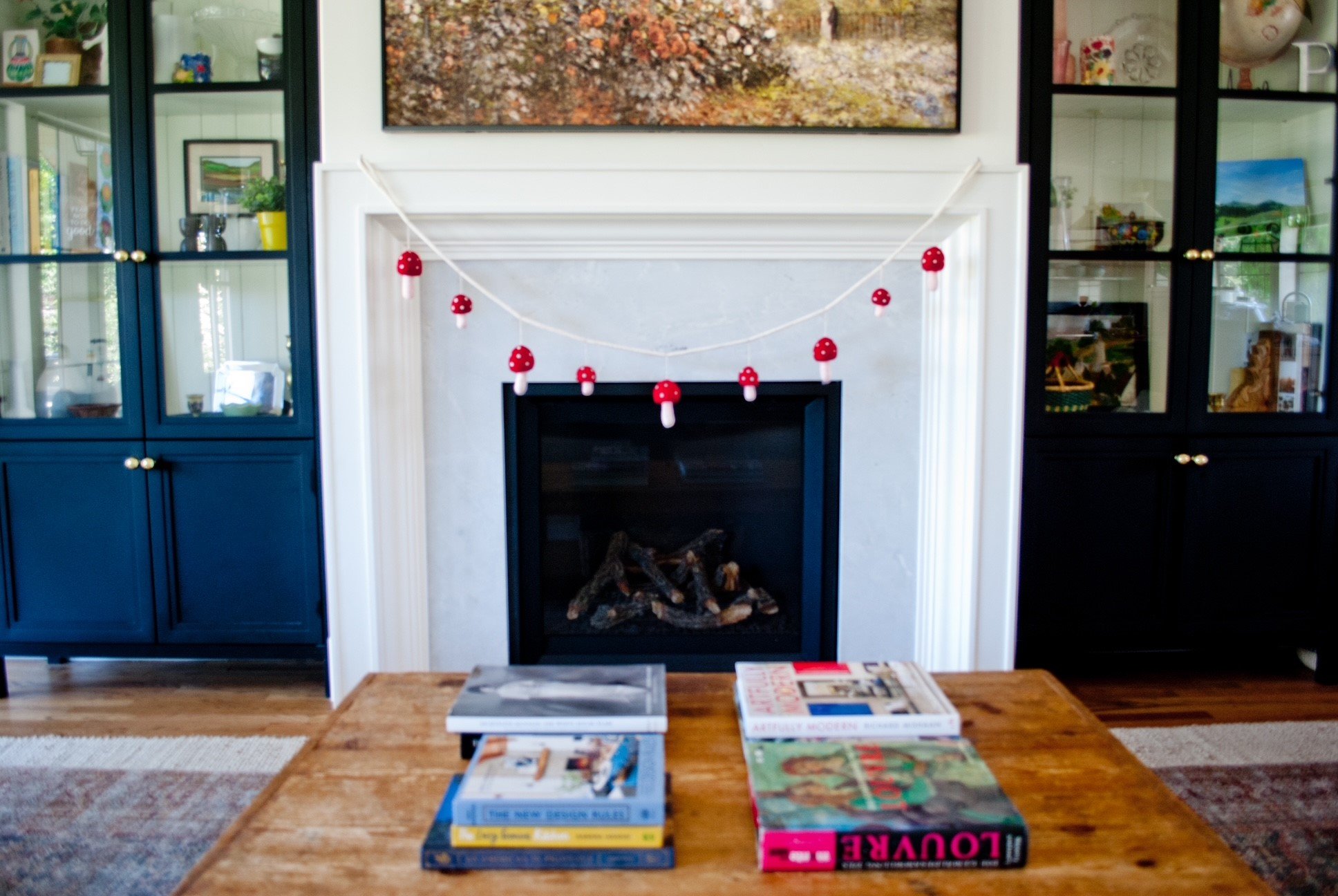 Living room with a white fireplace mantle decorated with a string of red mushroom-shaped ornaments. A wooden coffee table in front holds several magazines, and a framed landscape photo hangs above the fireplace. Flanking the fireplace are navy blue c