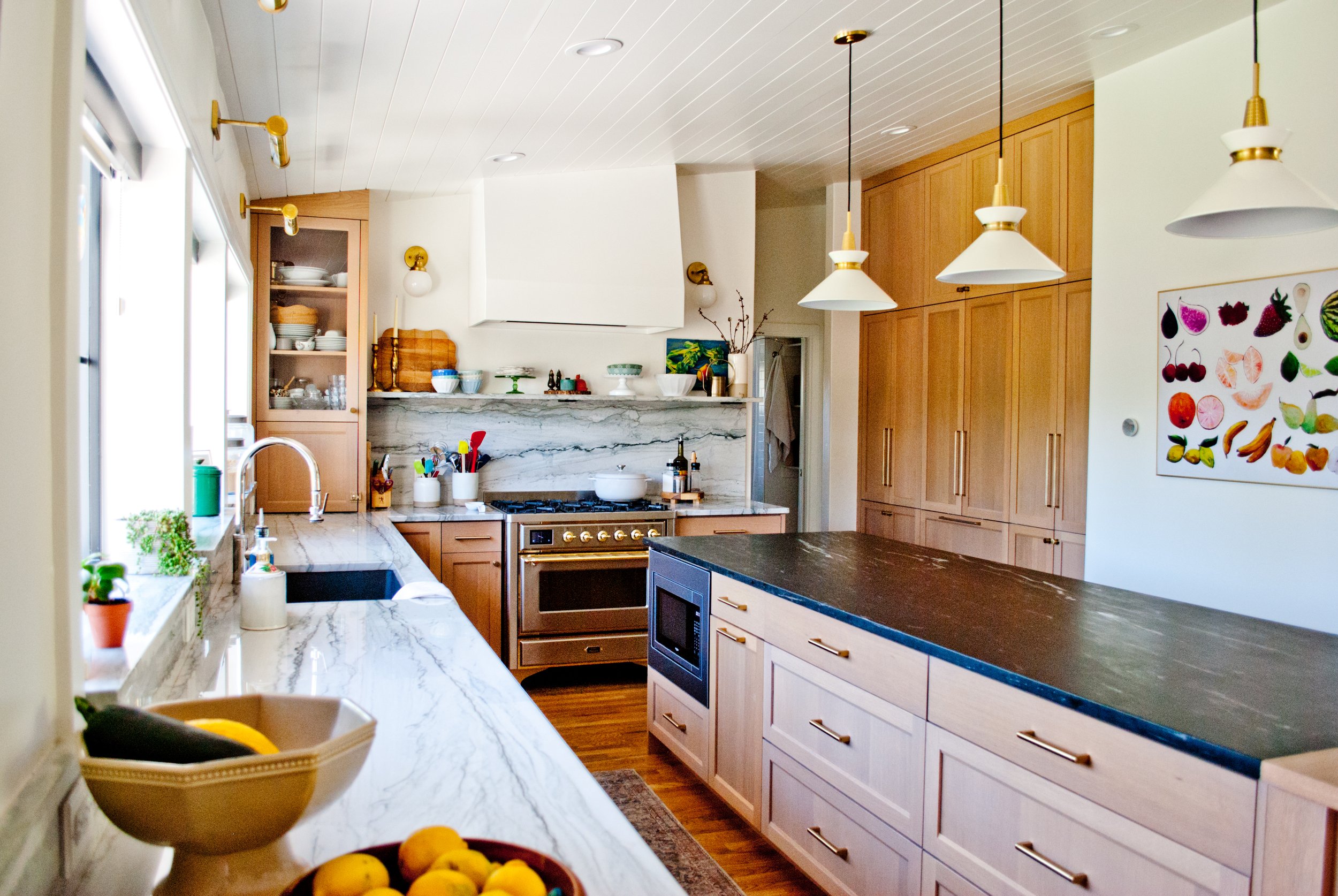 Modern kitchen with wooden cabinets, marble countertops, and a black-stone island. Features include hanging pendant lights, a marble backsplash, and a window with plants on the sill. Kitchenware and decorative items are organized on the countertops a