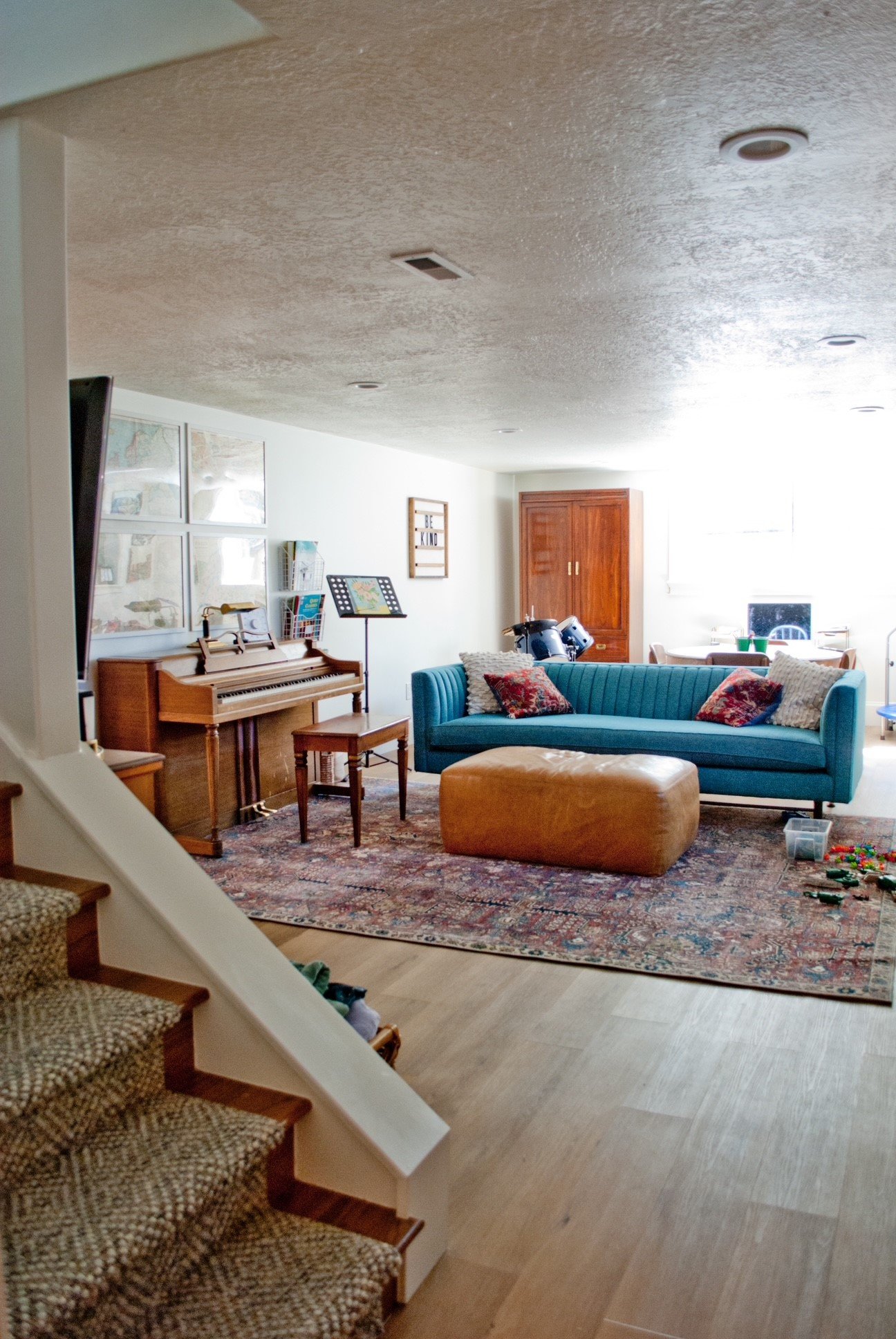 Living room with a blue sofa, brown ottoman, upright piano, and a patterned area rug near staircase.