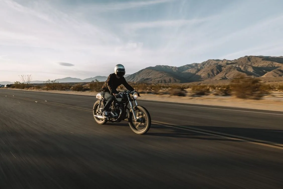 Motorcycle rider wearing a helmet riding down a paved road through a desert landscape with mountains in the background at sunset.