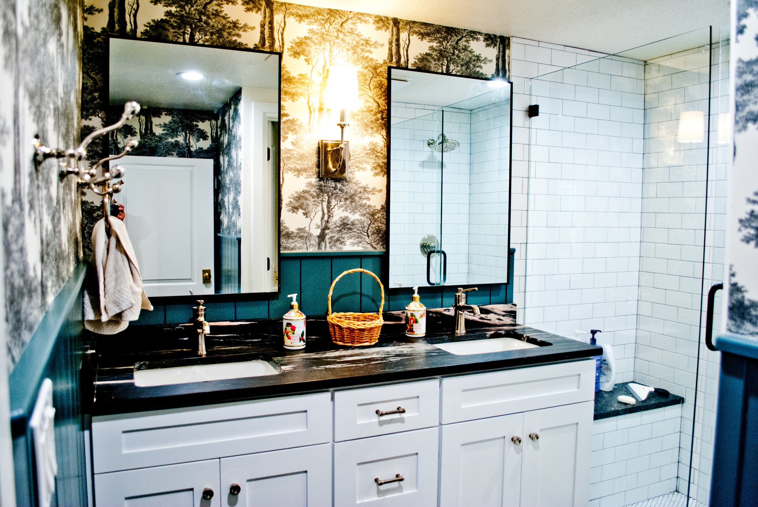 Bathroom with double sinks, large mirror, wallpaper with trees, and a walk-in shower with white subway tiles and a glass door.