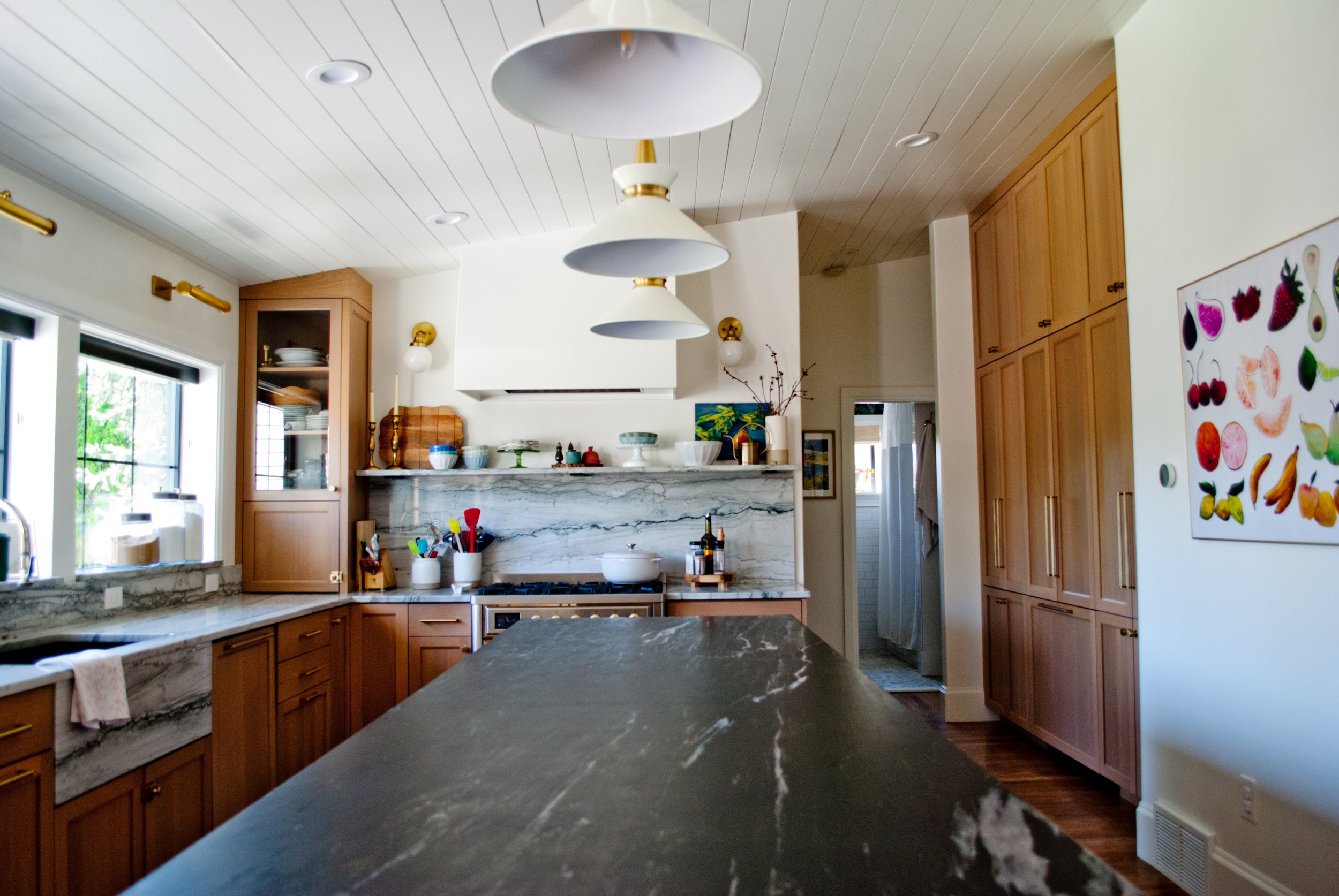 A kitchen with wooden cabinets, marble countertops, a modern pendant light fixture, and various kitchen items including bowls, utensils, and a fruit poster on the wall.