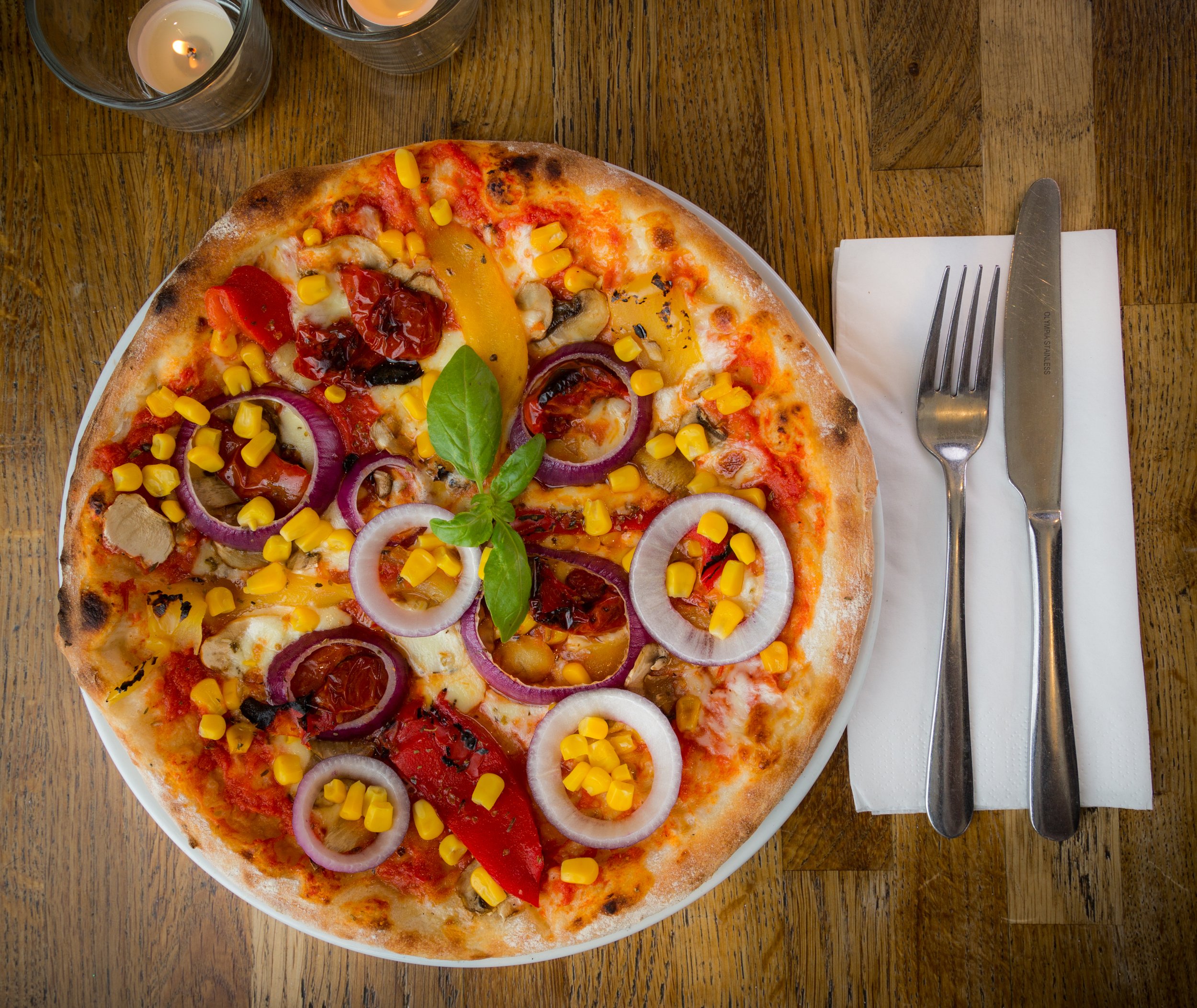 Pizza with red onions, sweet corn, bell peppers, and basil on a wooden table with utensils and candles.