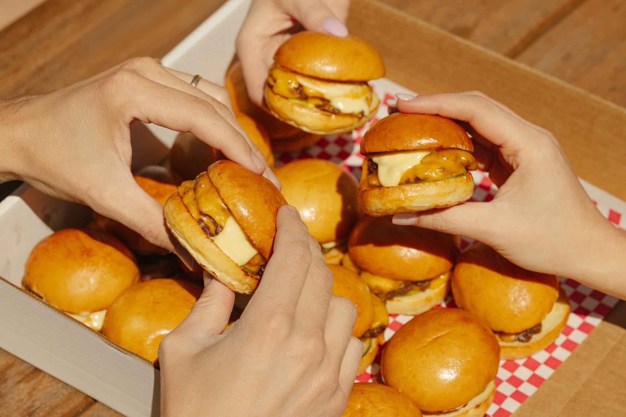 Hands reaching into a box of cheeseburger sliders