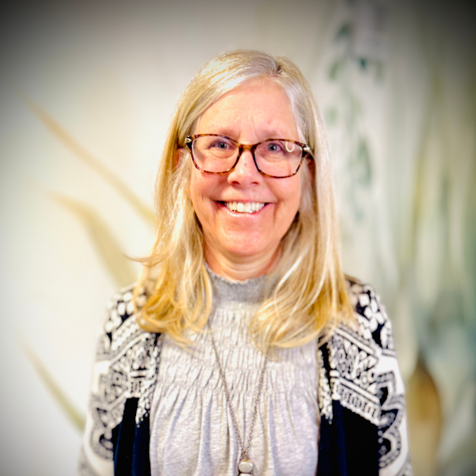 A smiling woman with blonde hair and glasses standing outdoors with green trees in the background.