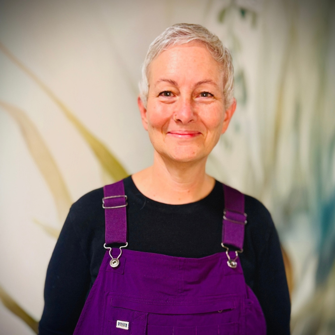 A woman with short gray hair smiling, wearing a black shirt and green overalls, standing in front of a bookshelf with some plants in the background.