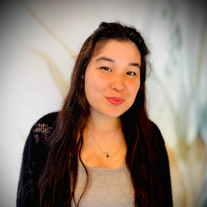 Smiling young woman with long dark hair, wearing a black and pink patterned top, in a room with bookshelves and plants in the background.