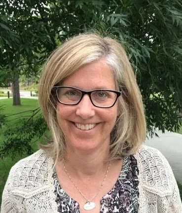 A smiling woman with blonde hair and glasses standing outdoors with green trees in the background.
