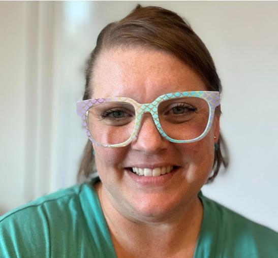A woman wearing large, colorful, patterned glasses and a teal shirt, smiling at the camera.