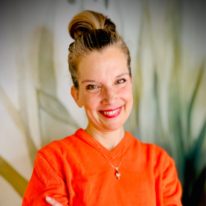 A woman with shoulder-length wavy hair wearing a pink sweater with red hearts, smiling in front of a bookshelf filled with books and a potted plant.