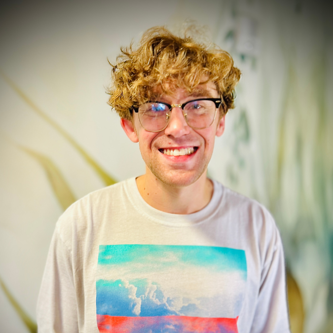 Young man with curly hair and glasses smiling in a room with bookshelf in the background.