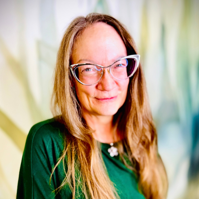 A woman with long wavy hair wearing glasses and a black knit sweater, smiling, in front of bookshelf background with plants.