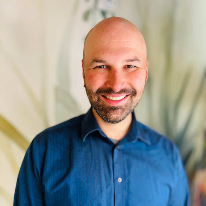 A smiling man with a beard, wearing earrings and a colorful patterned shirt, standing in front of a bookshelf filled with books.