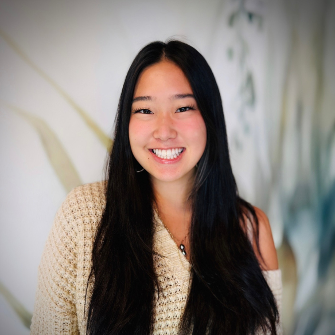 Young woman with long dark hair smiling indoors with books and plants in the background.