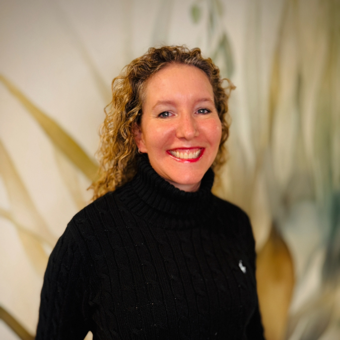 A woman with curly hair and glasses smiling, wearing a red jacket over a black top, standing in an indoor setting with shelves and plants in the background.