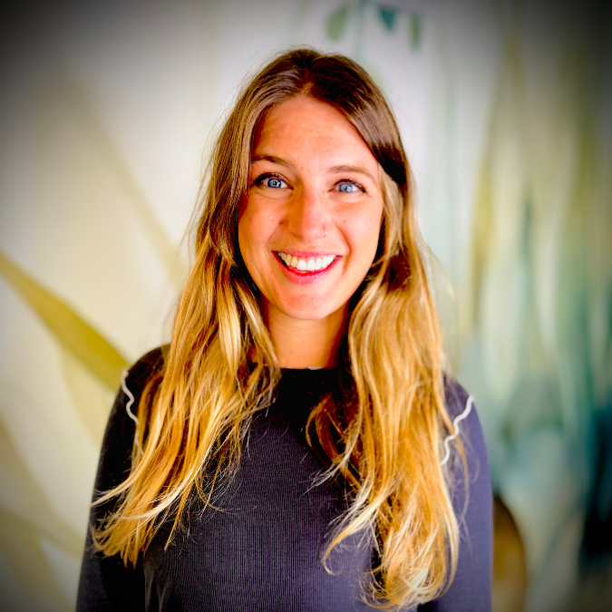 A young woman with long, wavy blonde hair smiling at the camera. Behind her, there is a bookshelf filled with colorful books and some potted plants, indicating an indoor setting, possibly a library or a study area.