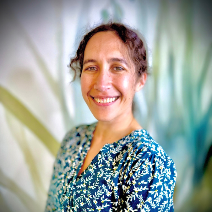 A woman with brown curly hair wearing a blue polka dot shirt, smiling in front of a bookshelf and green plants.