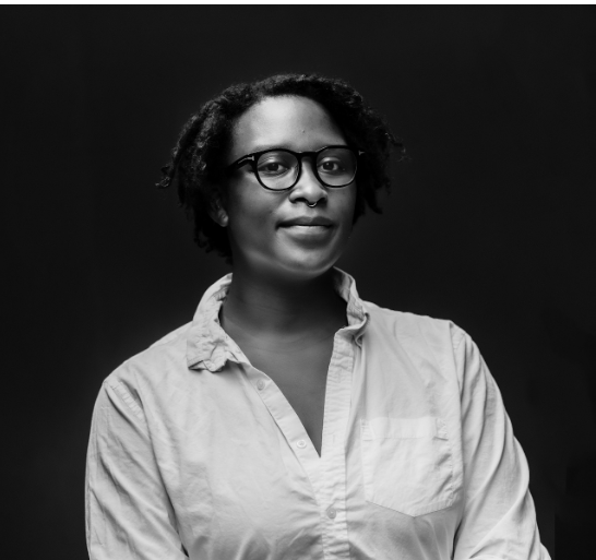 Portrait of a woman with glasses and short natural hair, wearing a light-colored button-up shirt, against a dark background.