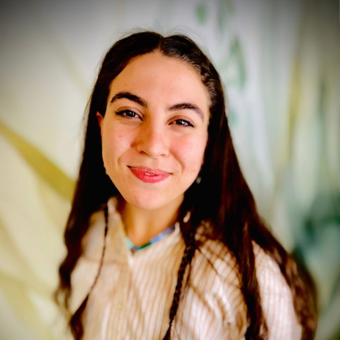 A young woman with long dark wavy hair smiling at camera, wearing a dark top, in a room with bookshelves and green plants in the background.