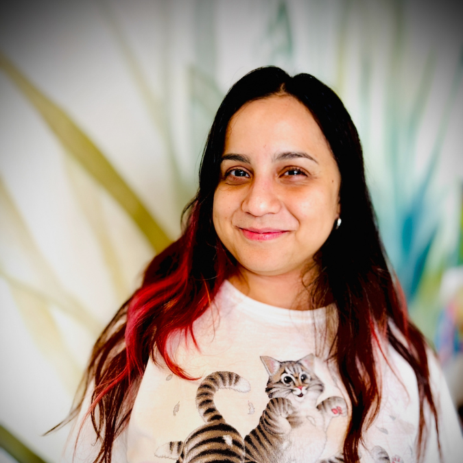 A woman with long, dark brown hair and light skin, smiling in front of a bookshelf with colorful items and books.