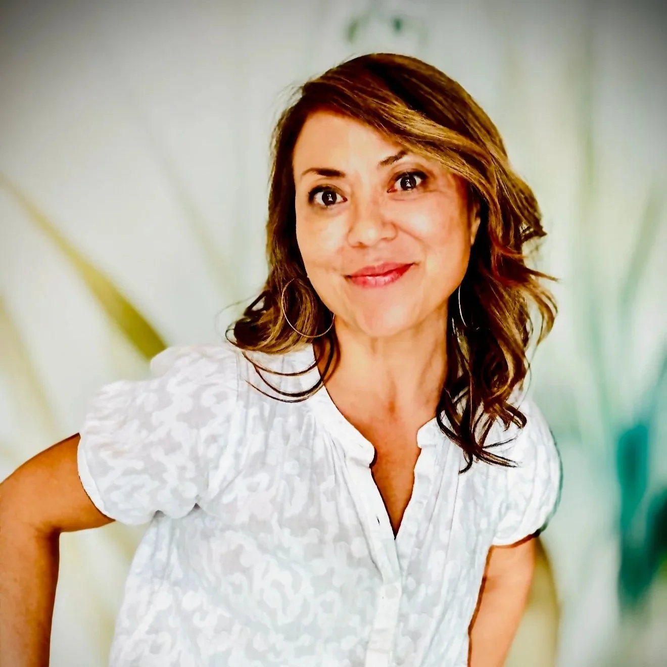 A woman with shoulder-length brown hair, wearing a red and white floral blouse, standing in front of a bookshelf with colorful binders and plants.