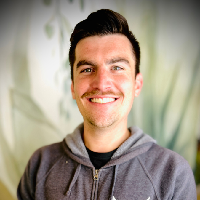 A smiling man with dark hair wearing a blue Adidas t-shirt, standing in front of a bookshelf and some plants.